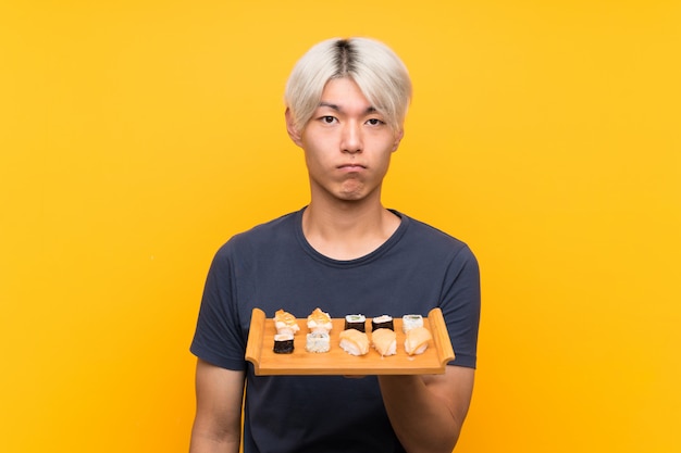 Young asian man with sushi over isolated yellow sad