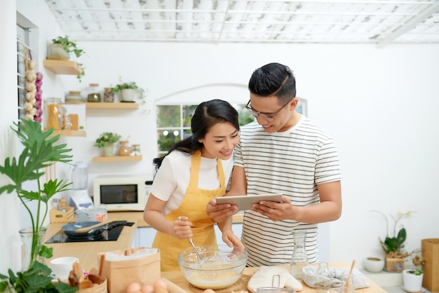 若いアジア人の男性と女性が一緒にケーキとパンを卵と一緒に調理し、家でリラックスして幸せな小麦粉のタブレットからメニューを探します