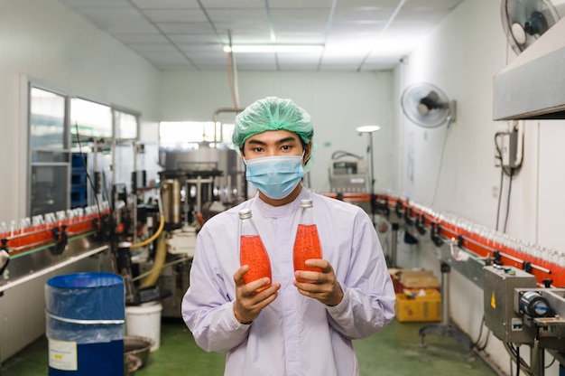 Young asian man worker in sterile uniform showing product of bottled red juice in production line at beverage processing factory