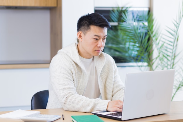 부엌에 있는 테이블에 앉아 노트북 컴퓨터로 집에서 일하는 젊은 아시아 남자