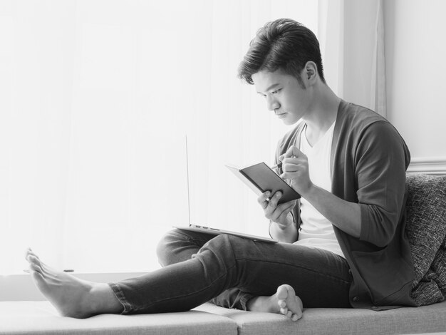 Young Asian man writing in a book when using laptop