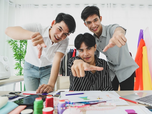 Giovani uomini asiatici che sorridono e puntano alla telecamera mentre lavorano insieme in uno studio di sartoria professionale