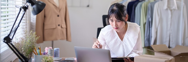 Foto giovani commercianti asiatiche che controllano gli ordini online e le informazioni dei clienti sul portatile per prendere appunti nel blocco mentre lavorano e spedire la consegna per l'attività di acquisto online in ufficio a casa