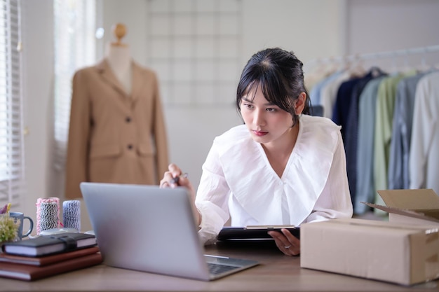 젊은 아시아 상인 여성은 노트북에서 온라인 주문 및 클라이언트 정보를 확인하고 작업하는 동안 클립보드에 메모를 취하고 홈 오피스에서 온라인 쇼핑 비즈니스를 위해 배송 배달을합니다.