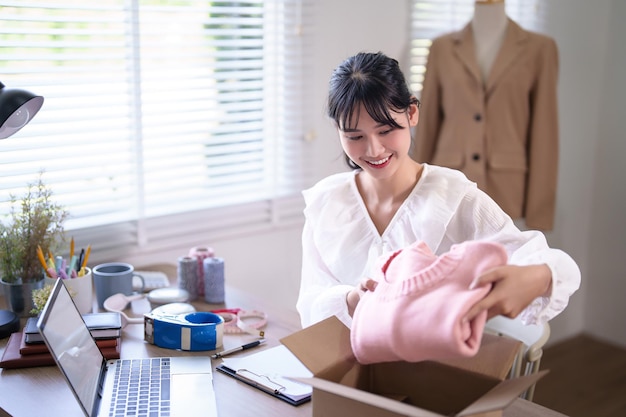 Foto giovani commercianti asiatiche che controllano l'acquisto online su laptop e imballano vestiti prodotto in pacco per la consegna di spedizione per il cliente mentre lavorano su online shopping business in ufficio a casa