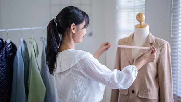 Photo young asian merchant women checking size details of new fashion clothes in mannequin with measuring tape while taking notes in clipboard and working about online shopping business in home office