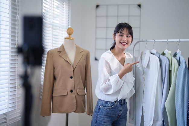 Photo young asian merchant women using smartphone to recording live streaming and presenting new clothes on rack to selling in social media while working and shipping delivery for online shopping business