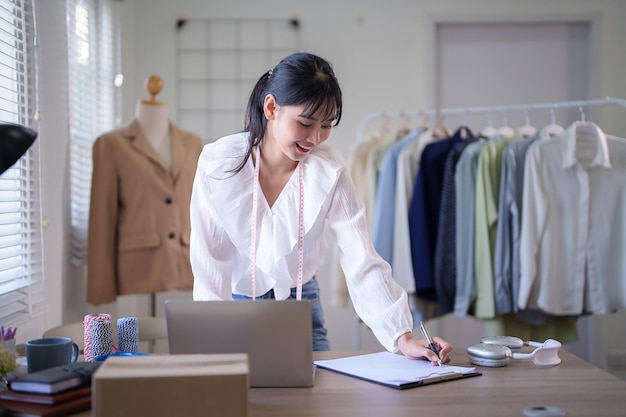 Foto giovane mercanti asiatiche che indossano nastro adesivo al collo e scrivono le informazioni dei clienti su carta mentre controllano gli ordini online sul portatile e lavorano sugli acquisti online