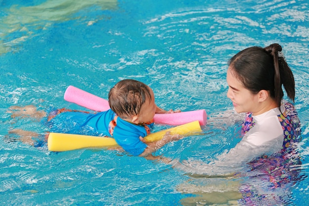 Young Asian mom teaching baby boy in swimming pool with noodle foam.