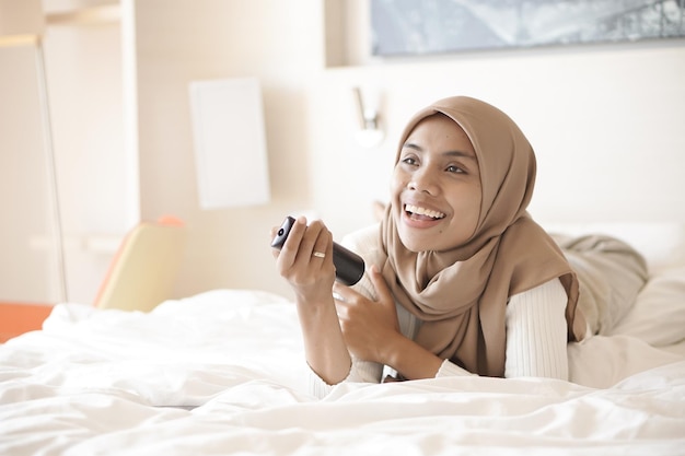 Young asian mother in bed cheerfully watching television in hotel room on vacation