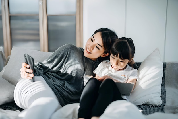 Young Asian mother and little daughter lying on the bed using smartphone and reading book they are having a relaxing time at home Beautiful sunlight illuminated them through window