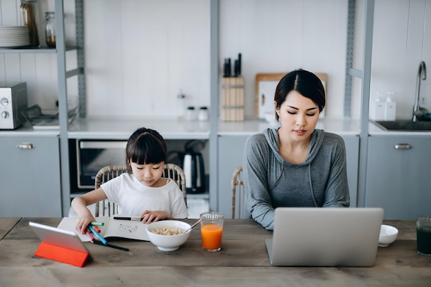 Young Asian mother working from home on a laptop while little daughter is studying from home She is attending online school classes with a digital tablet and doing homework at home