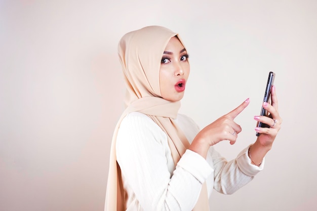 ヘッドスカーフを身に着けている若いアジアのイスラム教徒の美しい少女はスマートフォンにショックを受けています