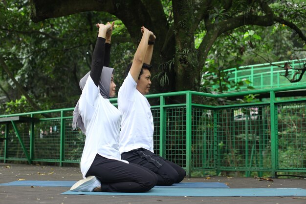 Young Asian muslim couple stretching and practicing yoga together on mattress at the park.