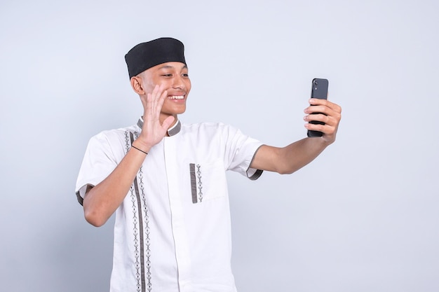 젊은 아시아 무슬림 남성이 스마트폰에서 비디오 통화나 라이브 스트리밍을 하면서 손을 흔들고 있다.
