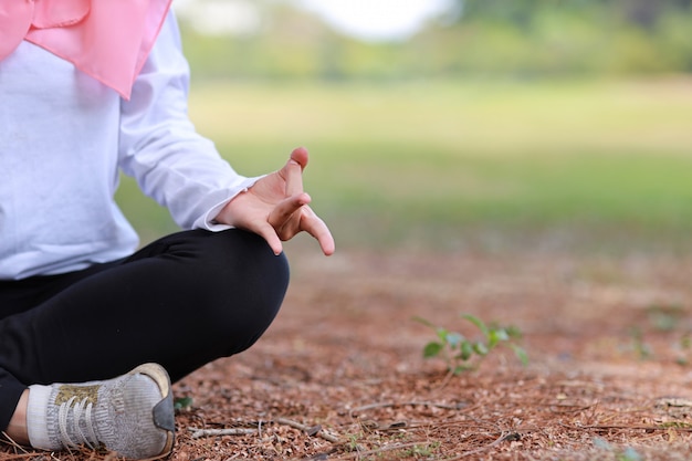 Foto giovane mano musulmana asiatica della donna che si siede sull'erba e che gode della meditazione. la bella ragazza in abiti sportivi con l'hijab rosa pratica l'yoga negli alberi di verde della natura con pace e calma. concetto sano e sportivo