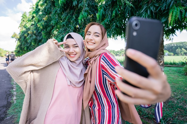 Young asian muslim woman in head scarf meet friends and using phone in the park for selfie or video calling