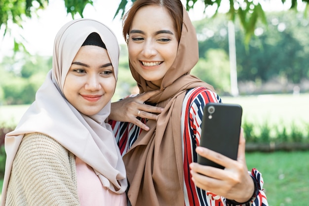 Young asian muslim woman in head scarf meet friends and using phone in the park for selfie or video calling