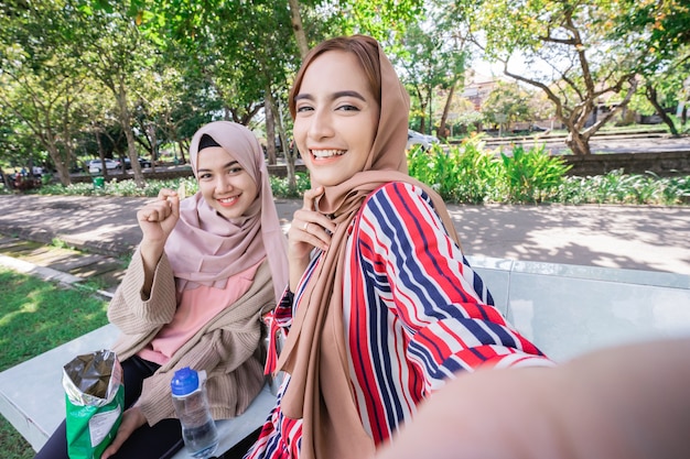 머리 스카프에 젊은 아시아 무슬림 여성이 친구를 만나고 공원에서 전화를 사용하여 함께 셀카를 찍습니다.