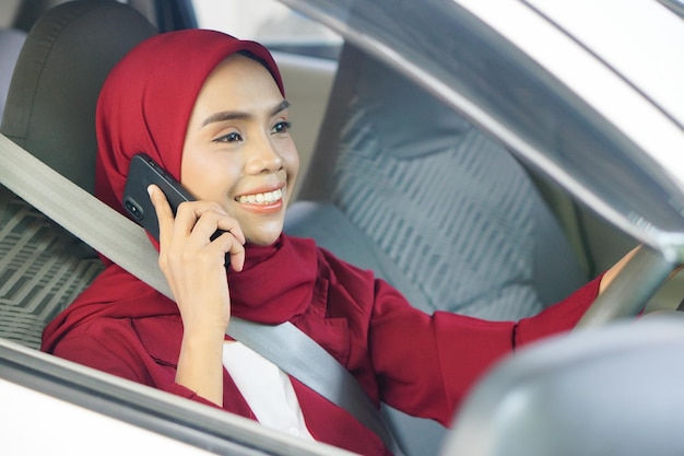 Молодая азиатская мусульманка в хиджабе с помощью смартфона улыбается в машине