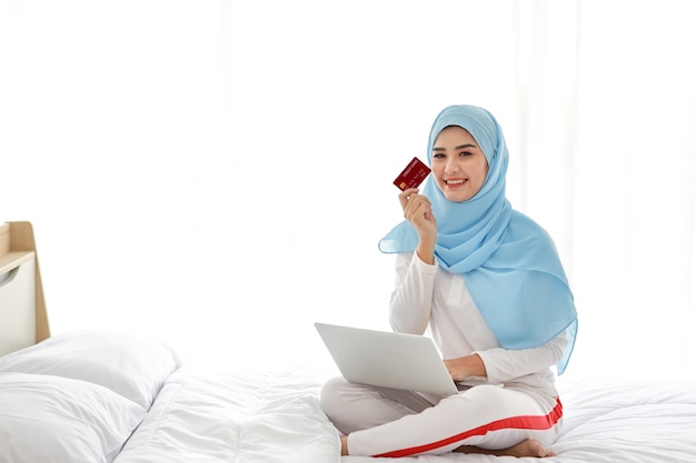 Молодая азиатская мусульманская женщина, сидящая на кровати и держащая кредитную карту с компьютером в спальне.