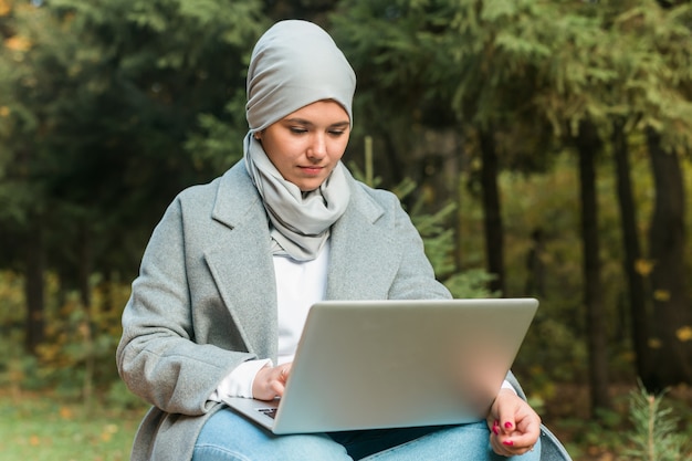 Young asian muslim woman using laptop at autumn park online education online shopping and social