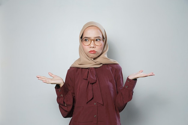 Молодая азиатка-мусульманка в очках, указывающая обеими улыбающимися руками, чувствует себя уверенно и счастливо