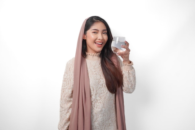 アジア系イスラム教徒の女性がヘッドスカーフを身に着けカメラに向かって微笑み白い背景で隔離された水のグラスを持っていますラマダンのコンセプト
