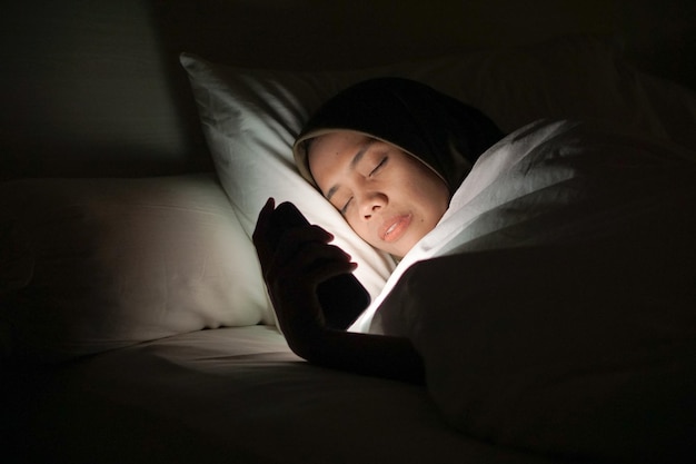 Young Asian Muslim woman wearing a headscarf while lying in bed and using a mobile phone at night