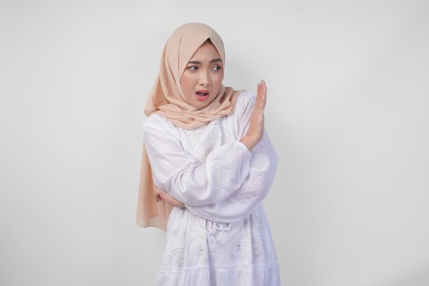 Молодая азиатская мусульманка в хиджабе и белом платье, делая жест отказа, отказывается использовать руки для копирования пространства с левой стороны, изолированного белым фоном, концепция Рамадана и Ид Мубарак