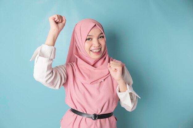 A young asian muslim woman with a happy successful expression wearing pink hijab isolated by blue background