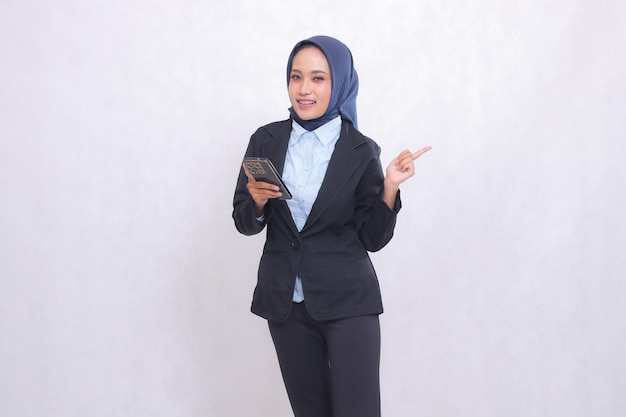 Young Asian office woman wearing a hijab stands happily at the camera and points to the left while c