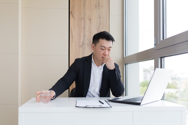 Young asian office worker suffering from headache at work in the office drinks medicine pill with water. Sick man in a suit at the computer indoors with severe pain ache uses the drug in workplace