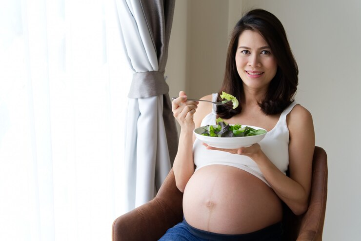 アジアの若い妊婦グリーンサラダと野菜を自宅の椅子に座っています。