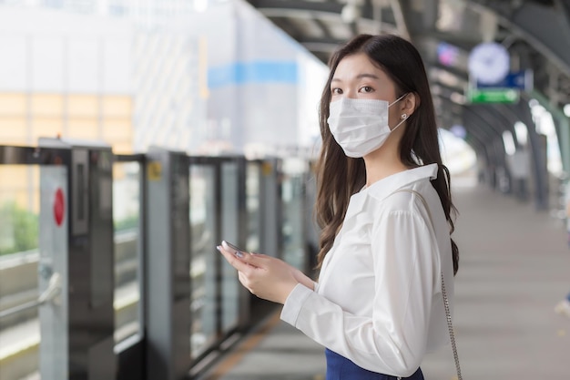 Una giovane donna d'affari asiatica professionale si trova su una stazione del treno sopraelevato in città indossando una maschera facciale