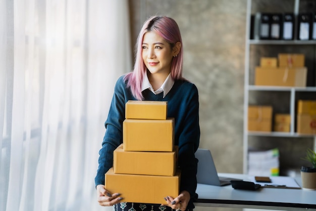 Young Asian SMEs woman holding parcels standing among several boxes in modern office