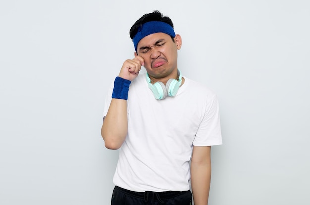 Young Asian sportman in blue headband and white tshirt with headphones rubs tears wants to cry feels depressed has a problem isolated on white background Workout sport concept