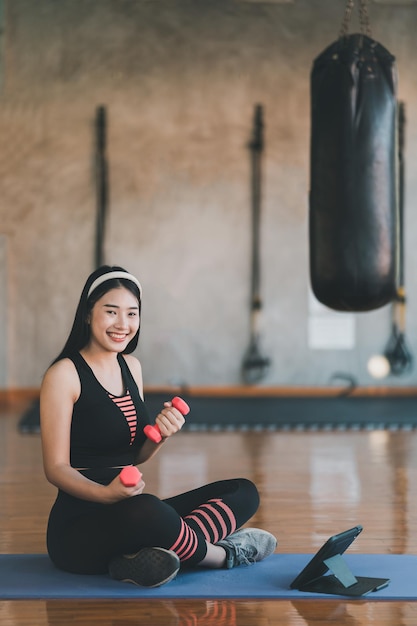 Молодая азиатская спортсменка сидит на коврике для йоги и делает упражнения с гантелями онлайн и смотрит потоковое видео на планшете в помещении в фитнес-зале Онлайн-тренировки по фитнесу из концепции тренажерного зала