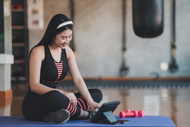 Young asian sportswoman sitting on yoga mat and doing exercise\
with dumbbell workout online and looking video streaming on tablet\
indoor at the fitness gym online fitness training from gym\
concept