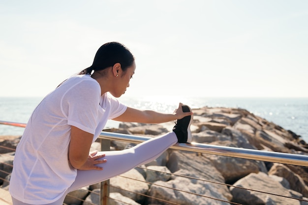 Молодая азиатская спортсменка, растягивающая ноги на берегу моря, концепция спорта и активного образа жизни