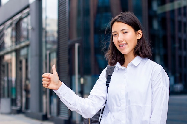 Молодая азиатская девушка студента показывая большой палец руки вверх и усмехаясь outdoors в белой рубашке с рюкзаком.