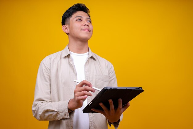 アジア系の若い男性学生がデジタルタブレットで働いて 上を見上げてアイデアを見つけています