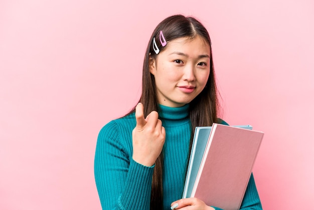 ピンクの背景に分離された本を持っている若いアジアの学生女性は、招待するようにあなたに指を指して近づいてくる