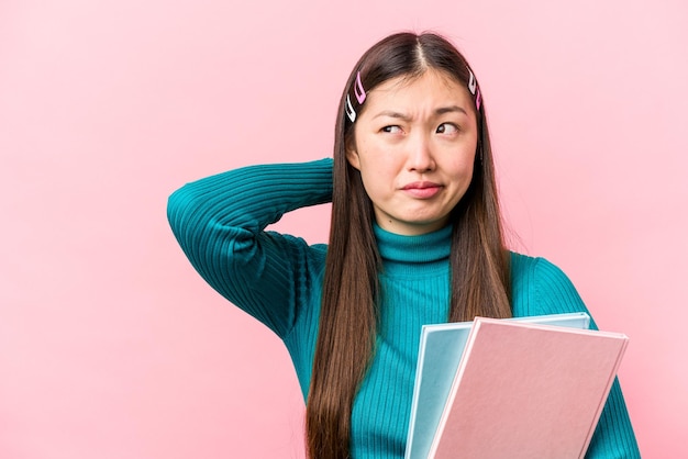 Photo young asian student woman holding books isolated on pink background touching back of head thinking and making a choice