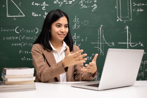 Young asian teacher woman sitting using laptop video conference\
with student female teacher training the mathematics in classroom\
from live stream with computer online course