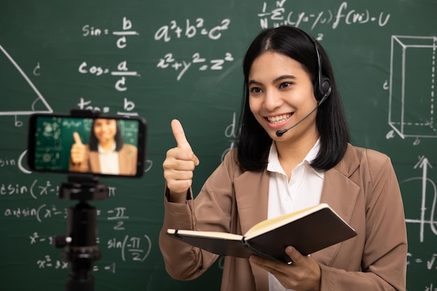 Young asian teacher woman standing looking at camera and video
conference with student female teacher training the mathematics in
classroom from live stream with smartphone online course