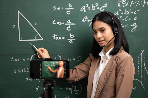 Giovane insegnante asiatica in piedi guardando la fotocamera e la videoconferenza con lo studente insegnante femminile che forma la matematica in classe dal live streaming con il corso online per smartphone