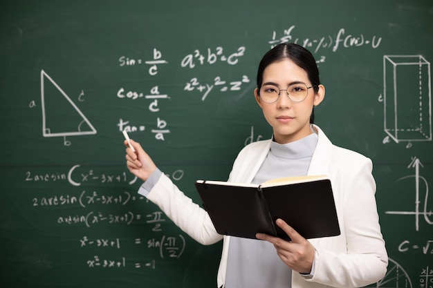 Young asian teacher woman teaching holding book and chalk video\
conference with student looking camera female teacher training the\
mathematics in classroom blackboard from online course