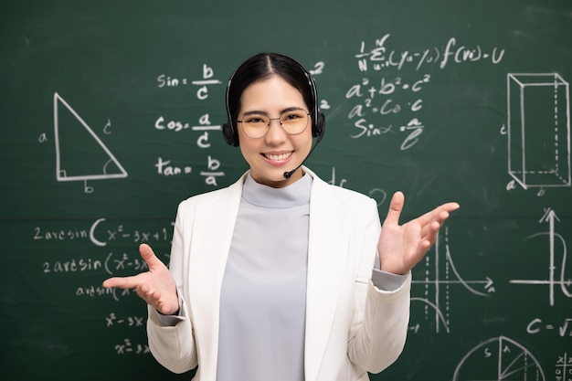 ヘッドセットを身に着けている若いアジアの教師の女性が学生とビデオ会議を話す教室の黒板オンラインコースで数学を訓練する女性教師