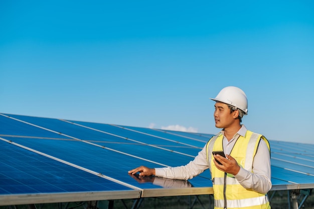 Young Asian technician man standing and talking on smartphone between long rows of photovoltaic solar panels copy space